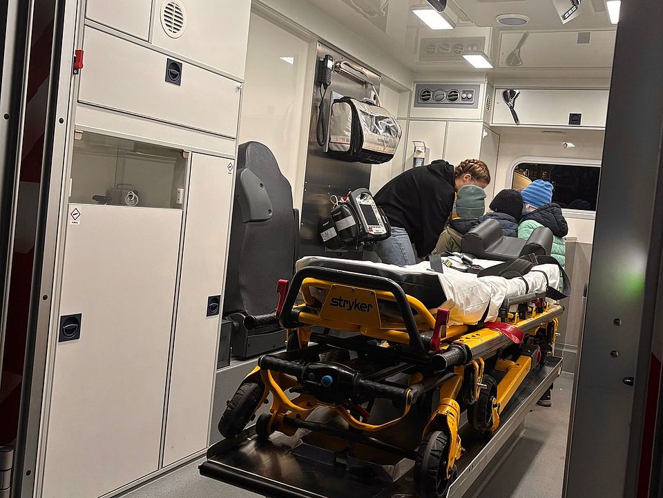2026_01_22_ Im RTW Man sieht ein Mitglied des JRK des DRK in einem Rettungswagen hinter der gelben Liege, mit drei Kindern. Die Ehrenamtliche zeigt den Kindern etwas, was aber nicht sichtbar ist