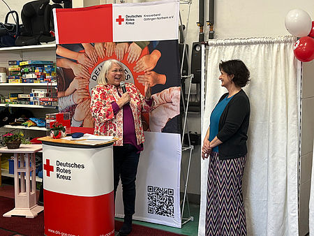 Sozialdezernentin der Stadt Göttingen, Anja Krause, steht am Redepult vor einem DRK‑Banner und spricht während der Einweihungsfeier des DRK‑Spendenzentrums Göttingen. Neben dem Redepult steht DRK-Kreisgeschäftsführerin Petra Reußner, dahinter Regale des Kaufhauses
