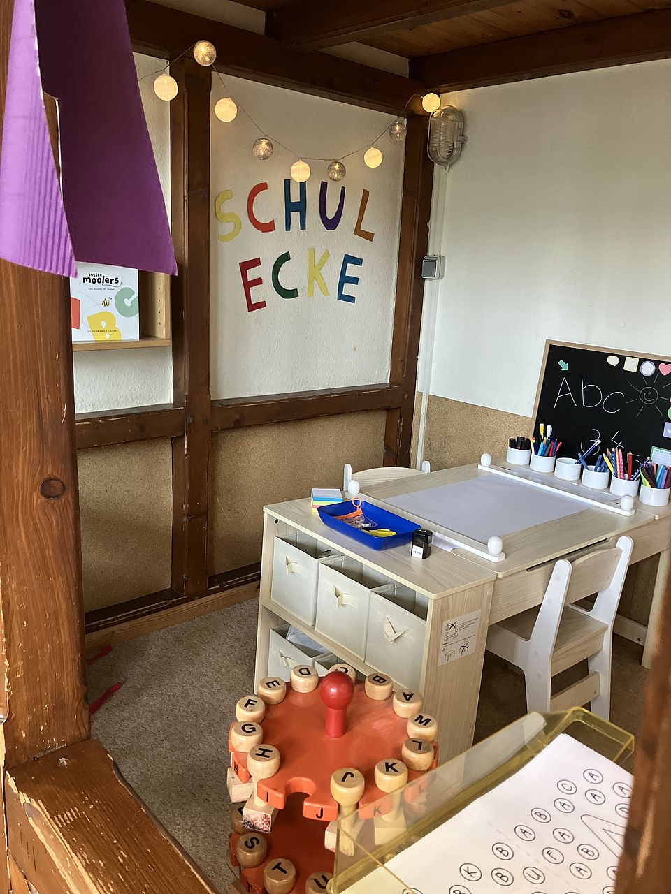 In einer Zimmerecke ist eine Schulecke mit Tisch, Tafel, Stiften und einem großen Stempel Ständer eingerichtet, in dem die Buchstaben von A-Z hängend aufbewahrt werden