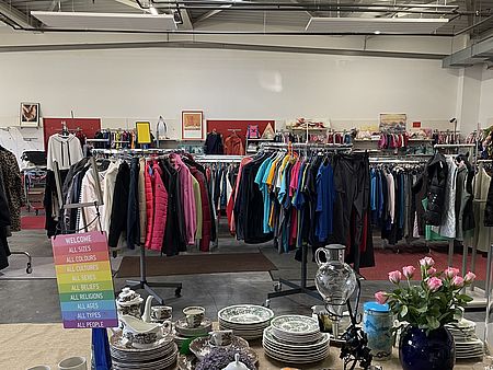 Innenraum des Göttinger Spendenzentrums des DRK-Kreisverbandes Göttingen‑Northeim e. V. Im Vordergrund steht ein Tisch mit verschiedenem Geschirr, Gläsern und dekorativen Gegenständen, darunter Tellerstapel, Tassen, eine Teekanne und ein Blumenstrauß in einer blauen Vase. Dahinter befinden sich mehrere Kleiderständer, dicht gefüllt mit bunter Kleidung wie Jacken, T‑Shirts und Pullovern. Der große Raum wirkt hell, offen und industriell, mit hohen Decken und sichtbaren Metallträgern