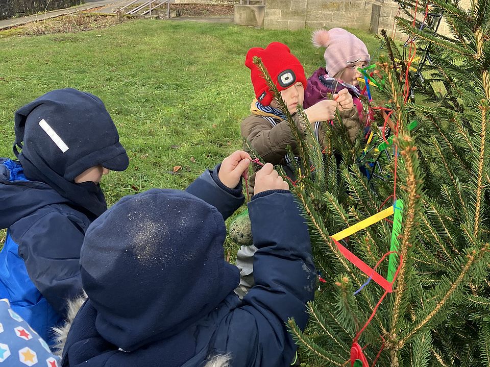 2025_12_11_Adventszeit_Kita_Wiershausen__3 Fünf Kinder schmücken einen Tannenbaum draußen bei Sonnenschein und binden bunte Bänder in die Zweige