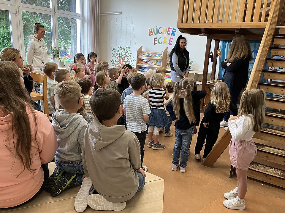 Eine Gruppe von Kita-Kindern stehen in Richtung einer Zimmerwand auf dem das Wort Bücherecke steht und auch Bücher im Regal zu sehen sind. Drei Kita-Mitarbeiterinnen stehen in der Gruppe und erklären etwas