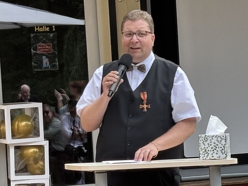 2026_07_04_Bundesverdienstkreuz_Jan_Störmer__4_.jpeg Jan Störmer mit Bundesverdienstkreuz spricht mit Mikrofon beim DRK-Kreisverband Göttingen-Northeim e. V. im Haus des Jugendrotkreuzes Einbeck