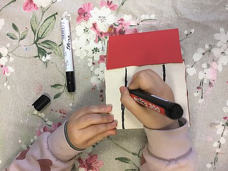 Zwei Kinderhände, die mit einem schwarzen Edding ein vorgebasteltes Haus aus Pappe mit Strichen bemalen