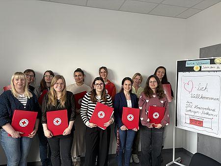 Gruppenfoto beim Willkommenstag des DRK-Kreisverbands Göttingen‑Northeim: Die Teilnehmenden stehen vor einem Whiteboard mit Willkommensbotschaft und halten rote DRK‑Mappen in den Händen