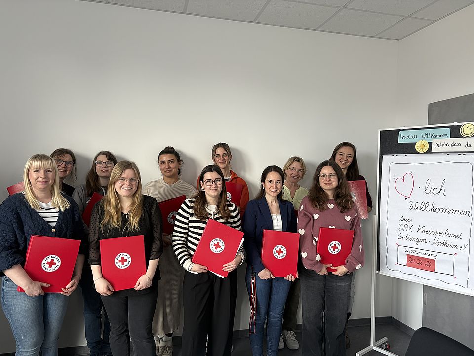 Gruppenfoto beim Willkommenstag des DRK-Kreisverbands Göttingen‑Northeim: Die Teilnehmenden stehen vor einem Whiteboard mit Willkommensbotschaft und halten rote DRK‑Mappen in den Händen
