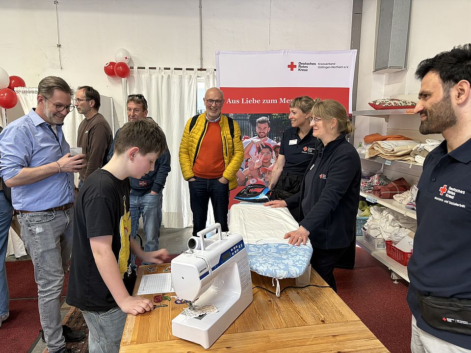 Im DRK‑Spendenzentrum Göttingen stehen Gäste und Mitarbeitende an einem Aktionstisch mit Nähstation. Auf dem Tisch stehen eine Nähmaschine und ein Bügelbrett, während mehrere Personen gemeinsam Stoffe ausprobieren und sich austauschen. Im Hintergrund sind ein DRK‑Banner mit der Aufschrift ‚Aus Liebe zum Menschen‘ sowie Regale mit Second‑Hand‑Artikeln zu sehen. Die Szene zeigt die belebte Nähecke der Einweihungsfeier am 3. März