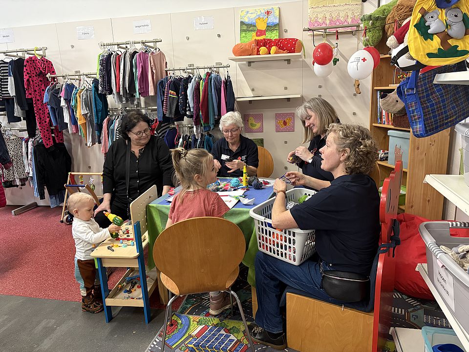 Kinder und Mitarbeitende sitzen an einem kleinen Tisch in der Kreativecke des neuen DRK‑Spendenzentrums Göttingen und basteln gemeinsam. Um sie herum stehen Regale mit Second‑Hand‑Kinderkleidung, Spielzeug und Dekoartikeln. Die Szene zeigt den familienfreundlichen Mitmachbereich der Einweihungsfeier am 3. März