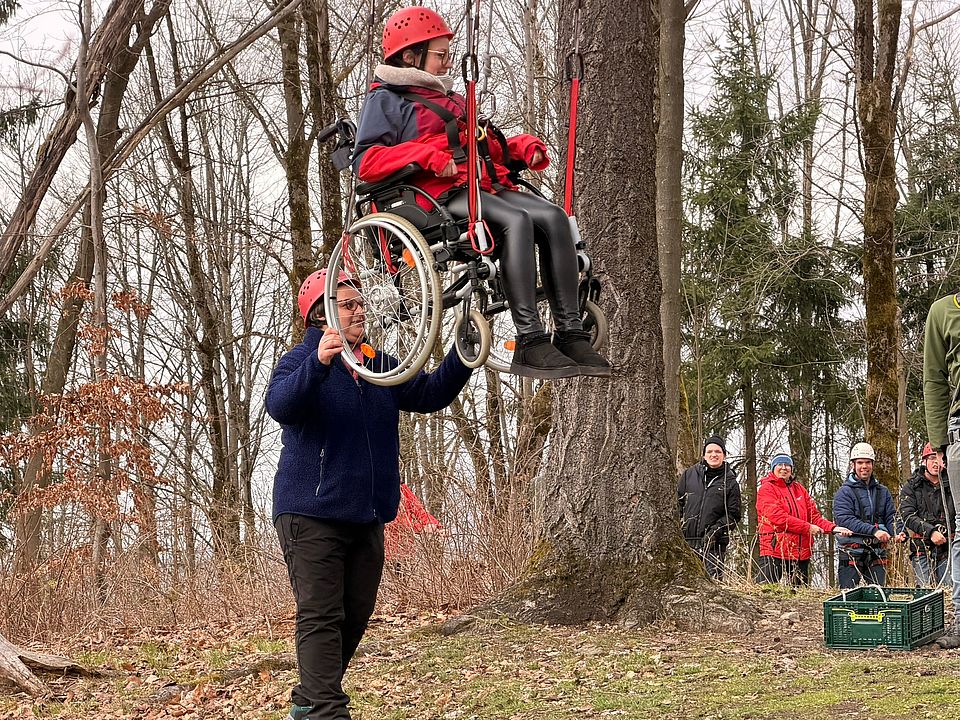 Rollstuhlschaukel im Wald bei der Ferienfreizeit des FED des DRK-Kreisverbandes Göttingen-Northeim e. V. Eine Teilnehmerin hängt mit Rollstuhl in der Luft, herum stehen andere Teilnehmende und eine Person hält den Rollstuhl fest
