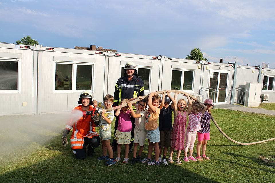 Acht Kita-Vorschul-Kinder halten einen Schlauch aus dem Wasser spritzt. Ein Feuerwehrmann kniet am Schlauchanfang und unterstützt, ein weiterer Feuerwehrmann steht hinter den Kindern. Alle stehen vor grauen Containern, in denen sich Feuerwehrräume befinden.