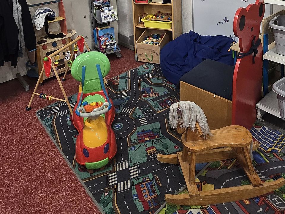 Kleine Spielecke im Innenraum des DRK-Spendenzentrums. Auf einem bunten Spielteppich mit Straßenmotiven stehen ein Holzschaukelpferd und ein farbiges Rutschfahrzeug. Links befinden sich Regale mit Kinderkleidung, Büchern und Spielzeug. Rechts stehen ein Sitzsack und ein roter Kinderstuhl in Tierform. Ein hohes Regal an der Wand ist mit Stofftieren und weiteren Spielsachen gefüllt. An der Wand hängt ein Schild mit der Aufschrift ‚Spielecke‘.