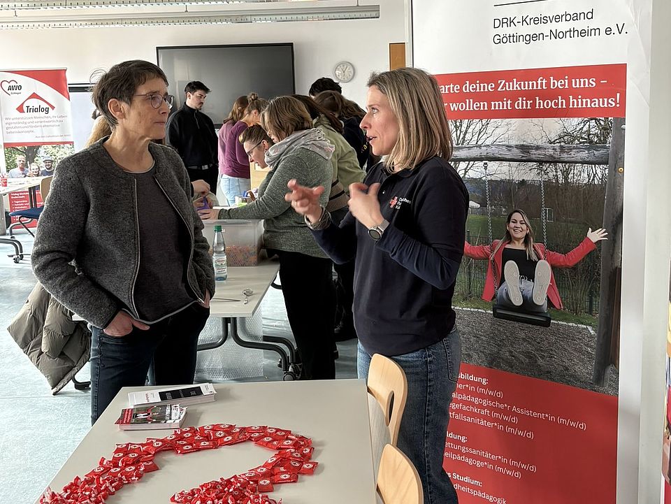 2026_02_25_Messe_Durchstarten_in_der_Sozialpägagogik_VHS__15_.JPEG In einem hellen Raum der VHS Göttingen Osterode stehen mehrere Personen an einem Infostand des Deutschen Roten Kreuzes (DRK‑Kreisverband Göttingen‑Northeim e. V.) auf der Jobmesse „Durchstarten in der Sozialpädagogik“. Im Vordergrund befindet sich ein Tisch mit Informationsmaterialien und zahlreichen roten Kunststoffherzen, die zu einer großen Herzform ausgelegt sind. Rechts neben dem Tisch steht ein großes DRK‑Roll‑up‑Banner mit dem Schriftzug „Deutsches Rotes Kreuz“ und einem Motiv, das junge Menschen bei pädagogischen Aktivitäten zeigt. Eine Person in DRK‑Dienstkleidung spricht mit einer besuchenden Person, während im Hintergrund mehrere weitere Besucherinnen und Besucher an anderen Ständen Informationen einholen. Der Raum ist mit großen Fenstern und modernen Lichtleisten ausgestattet und wirkt gut gefüllt.