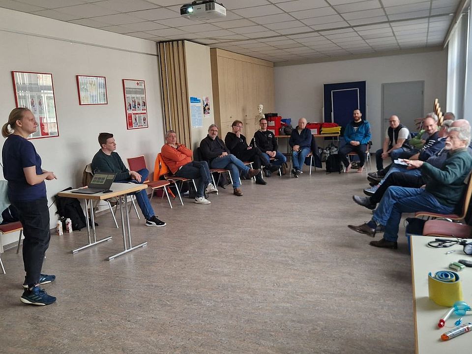 Eine Gruppe von Kursteilnehmenden sitzt in einem Schulungsraum in einem großen Stuhlkreis. Vorne steht eine Person neben einem Tisch mit Laptop und hält eine theoretische Unterrichtseinheit ab. Auf einem Seitentisch liegen verschiedene medizinische Übungsmaterialien bereit.