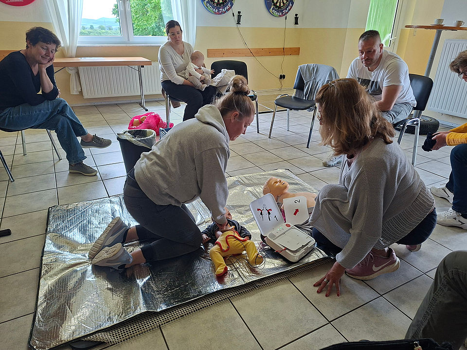 Zwei Teilnehmende am Erste-Hilfe Kurs des DRK knien auf dem Boden und üben die Wiederbelebung an einer Kinderpuppe mit Trainings-Defibrillator. Weitere Kursteilnehmende beobachten die Übung im Hintergrund.