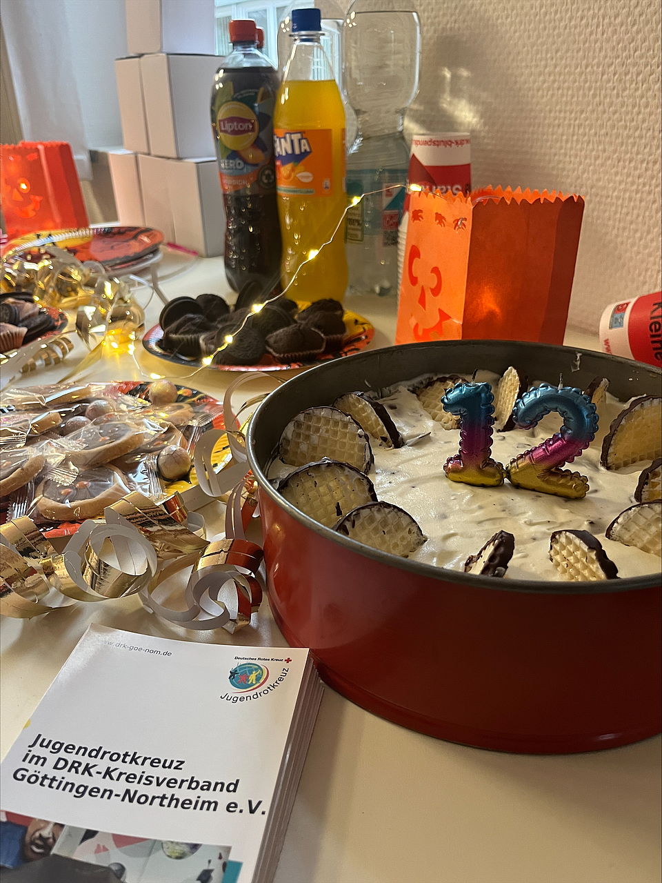 Eine Torte mit einer regenbogenfarbenen 12 steht auf einem gedeckten Tisch mit Süßigkeuten, Getränken, Luftschlangen, Lichterkette und JRK-Flyeren