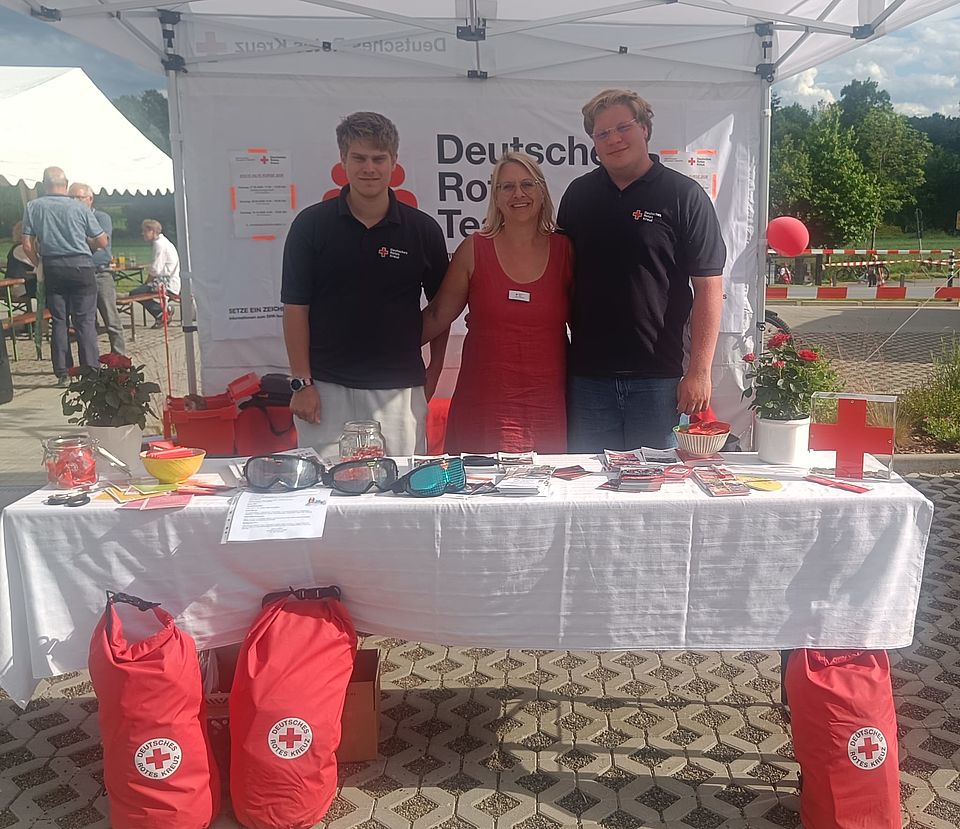 2026_06_06_1._Gesundheitstag_Ebergötzen_mit_OV_und_Erste_Hilfe__39_.jpg Tina Reich und Lars Trebing und eine weitere Person am DRK‑Infostand beim Gesundheitstag Ebergötzen mit Informationsmaterial und Notfallrucksäcken