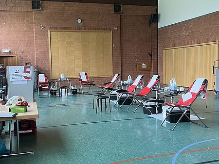 Das Bild zeigt eine Turnhalle mit fünf roten Stühlen, auf denen man für eine Blutspende liegen kann. Das Bild wurde bei einer Blutspende vom DRK-Ortsverein aufgenommen. Links sieht man Tische und Equipment für die Blutspende vom DRK