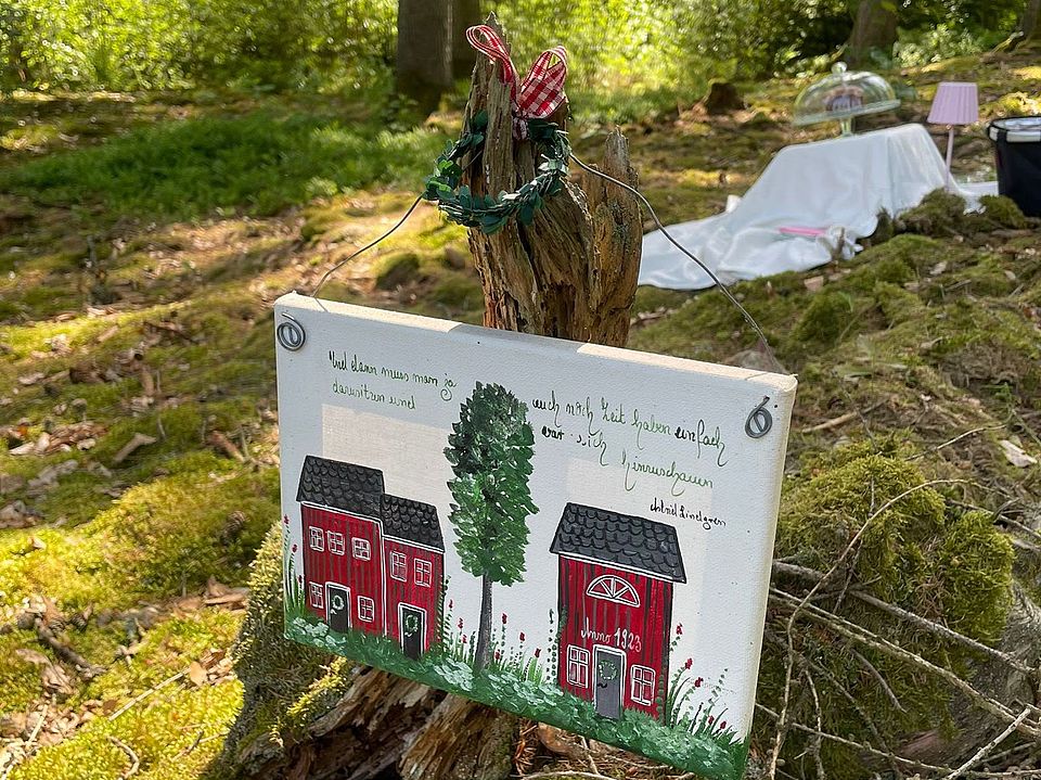 2026_05_26_Teamtag_Kita_Bühren__9_.jpeg Ein dekorierter Baumstumpf trägt ein handgemaltes Schild mit roten Häusern; im Hintergrund stehen Tisch, Decke und kleine Lampe zwischen Bäumen im Sonnenlicht.