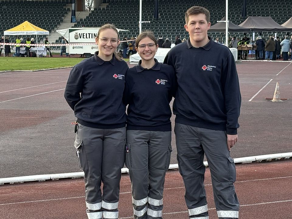 Drei Mitglieder der Bereitschaft des DRK Göttingen-Northeim e. V. stehen nebeneinander und lachen in die Kamera. Sie stehen im Jahnstadion Göttingen auf der Tartanbahn