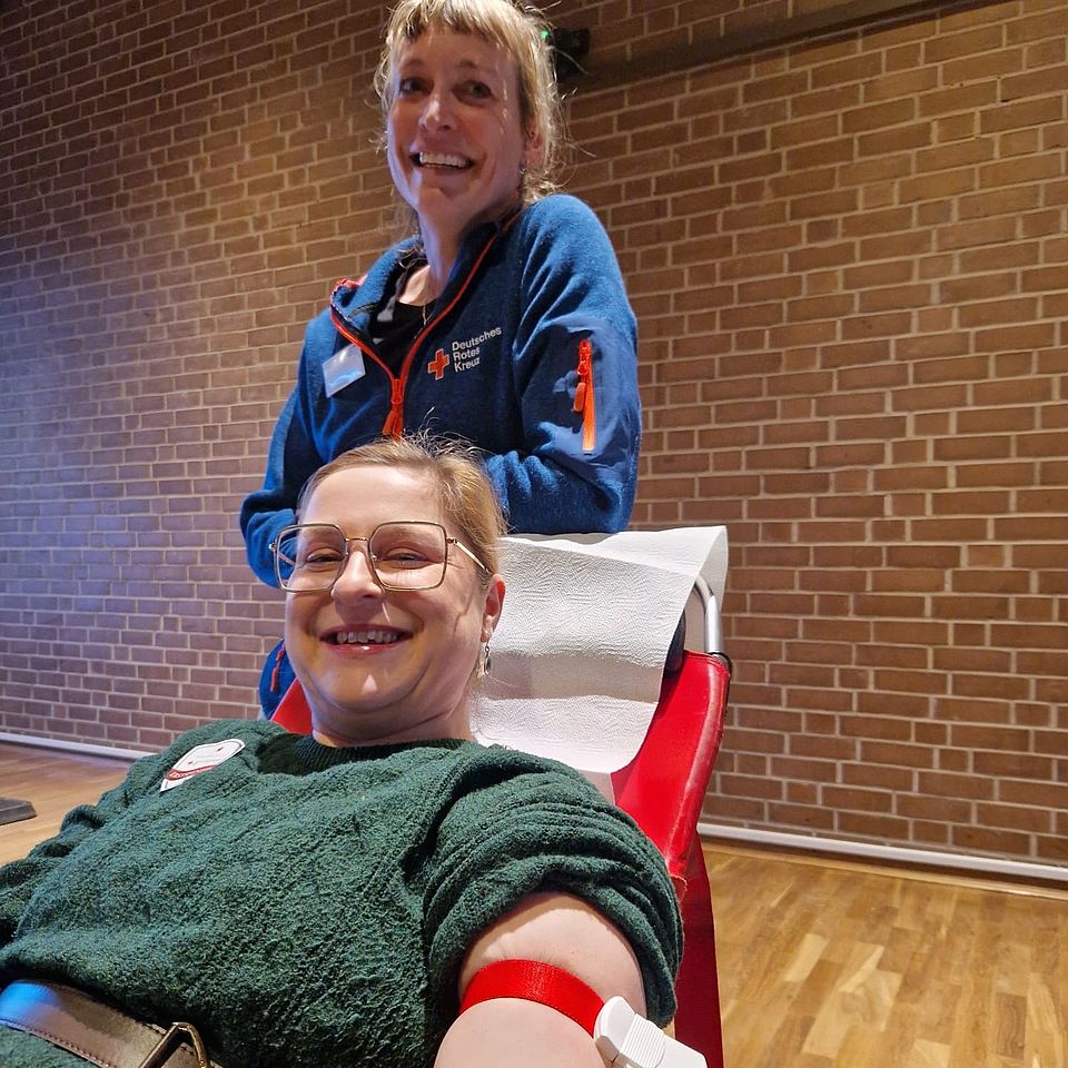 2026_02_Blutspende_Moringen__2_.jpeg In einem Raum mit heller Holzfußbodenfläche und einer Wand aus roten Ziegelsteinen liegt eine Person auf einer gepolsterten Blutspendeliege. Der angewinkelte Arm der Person ist mit einem roten Spendenband fixiert und an ein medizinisches Schlauchsystem angeschlossen. Hinter der liegenden Person steht eine weitere Person in einer Arbeitsjacke mit DRK‑Logo und Namensschild, die die Blutspende begleitet. Die Szene zeigt einen Moment während einer Blutspendeaktion.