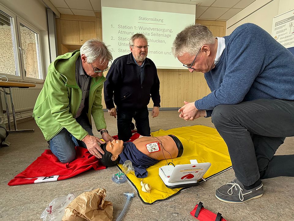 Teilnehmende eines Erste-Hilfe-Kurses üben die Versorgung einer lebensechten Reanimationspuppe. Zwei Personen knien auf dem Boden und prüfen Atemwege sowie Brustkorb, daneben steht ein automatisierter externer Defibrillator (AED) bereit. Im Hintergrund ist eine Präsentationsfolie mit Übungsstationen zu sehen.