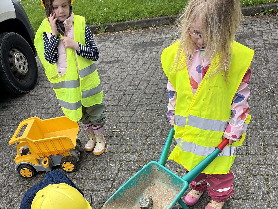 2026_05_27_Baustellentag_für_die_Kinder__4_.jpeg Ein Kind schiebt eine kleine Schubkarre mit Sand und Steinen, während ein weiteres Steine aus einem Eimer nimmt. Beide tragen Warnwesten und spielen das Arbeiten auf einer Baustelle nach.
