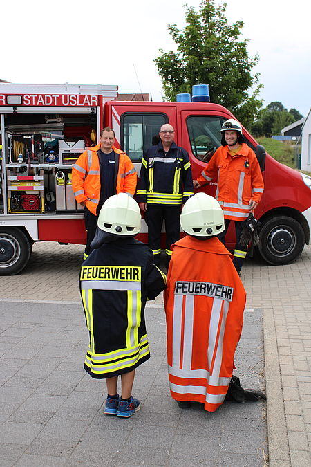 Zwei Kita-Kinder stehen mit Feuerwehrjacken und Helmen drei Feuerwehrmännern gegenüber. Diese stehen vor einem geöffneten Feuerwehrauto und schauen die Kinder an. Die Kinder sind mit dem Rücken zu sehen, so dass der Schriftzug "Feuerwehr" auf den Jacken zu lesen ist.