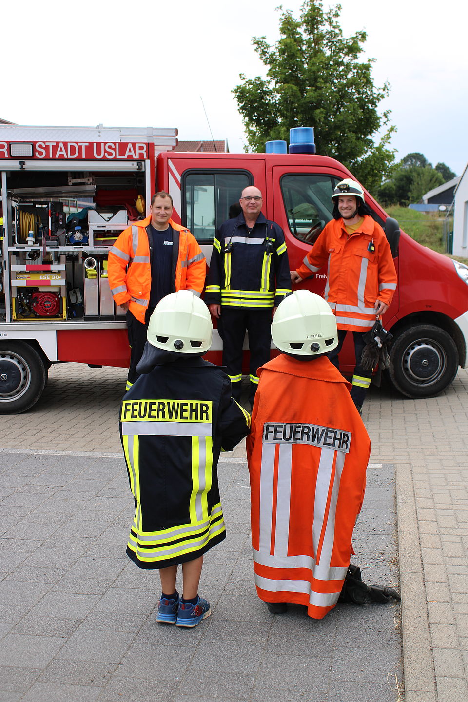 Zwei Kita-Kinder stehen mit Feuerwehrjacken und Helmen drei Feuerwehrmännern gegenüber. Diese stehen vor einem geöffneten Feuerwehrauto und schauen die Kinder an. Die Kinder sind mit dem Rücken zu sehen, so dass der Schriftzug "Feuerwehr" auf den Jacken zu lesen ist.