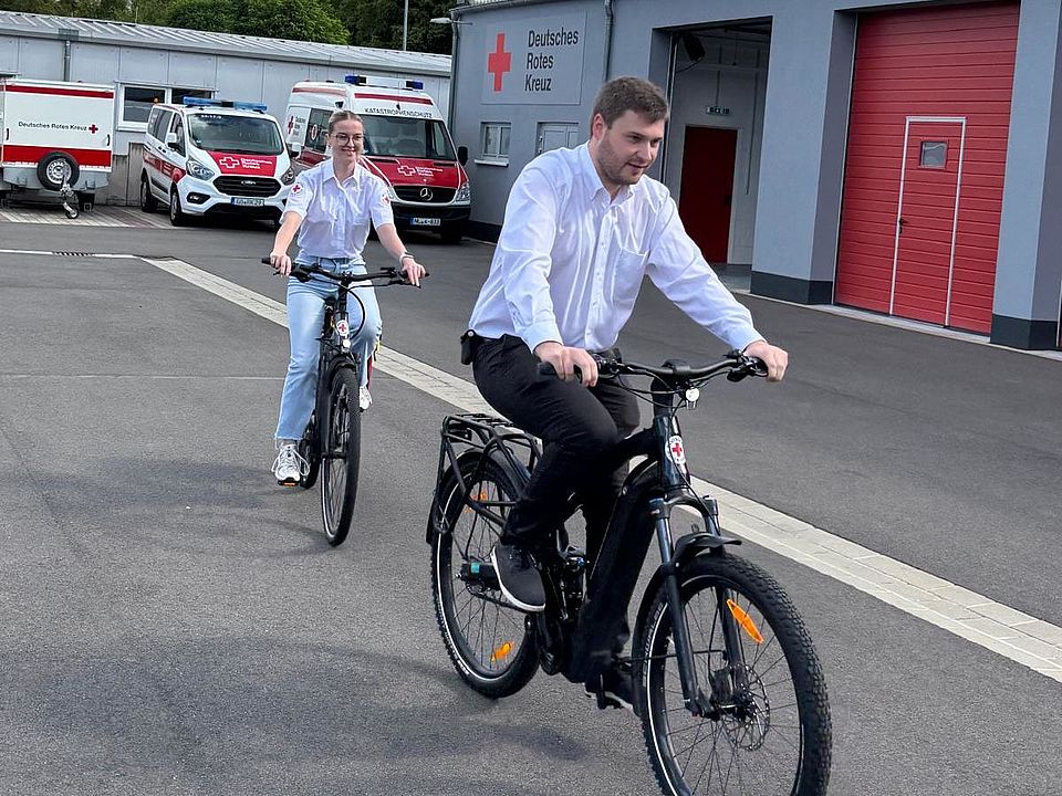 2026_08_11_Fahrradstaffel Gründung_Gutes_Foto_Michelle___Mika_auf_Rädern___DRK.jpeg Michelle Jonson und Mika Wolfram aus der Bereitschaftsleitung Göttingen testen die neuen E-Bikes der erstmals gegründeten Fahrradstaffel des DRK-Kreisverbands Göttingen-Northeim. Im Hintergrund sind Fahrzeuge des Rettungsdienstes und des Katastrophenschutzes sowie die Fahrzeughalle des DRK zu sehen.