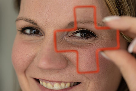 Augen eines Frauengesichtes. Vor ein Auge wird eine Büroklammer in Form eines Kreuzes gehalten, durch die die Frau lächelnd hindurchschaut