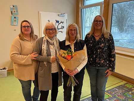 In einem freundlich gestalteten Raum einer Kindertagesstätte stehen vier Mitarbeiterinnen des DRK‑Kreisverbandes Göttingen‑Northeim e. V. eng beieinander und posieren für ein Gruppenfoto. Auf dem Bild zu sehen sind Sina Rohrig (Leitung der DRK‑Kita Jühnde), Uta Colburn (ehemalige Kita‑Fachberatung bis 2021), Gundel Matern (pädagogische Assistenzkraft) und Jenny Treichel (DRK‑Fachberatung Kita). Gundel Matern hält einen großen Blumenstrauß mit orangefarbenen und gelben Blüten in den Händen. Der Strauß symbolisiert ihre Verabschiedung in die Altersrente, die im Rahmen dieses Zusammentreffens gewürdigt wird. Der helle Raum ist mit kindgerechter Dekoration und einem Bild mit Vogelmotiven ausgestattet. Durch große Fenster fällt Tageslicht ein, draußen ist eine verschneite Landschaft zu erkennen. Auf dem Boden liegen farbenfrohe Spielteppiche. Die Szene vermittelt eine warme und wertschätzende Atmosphäre.