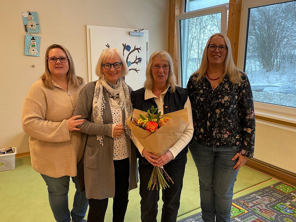 In einem freundlich gestalteten Raum einer Kindertagesstätte stehen vier Mitarbeiterinnen des DRK‑Kreisverbandes Göttingen‑Northeim e. V. eng beieinander und posieren für ein Gruppenfoto. Auf dem Bild zu sehen sind Sina Rohrig (Leitung der DRK‑Kita Jühnde), Uta Colburn (ehemalige Kita‑Fachberatung bis 2021), Gundel Matern (pädagogische Assistenzkraft) und Jenny Treichel (DRK‑Fachberatung Kita). Gundel Matern hält einen großen Blumenstrauß mit orangefarbenen und gelben Blüten in den Händen. Der Strauß symbolisiert ihre Verabschiedung in die Altersrente, die im Rahmen dieses Zusammentreffens gewürdigt wird. Der helle Raum ist mit kindgerechter Dekoration und einem Bild mit Vogelmotiven ausgestattet. Durch große Fenster fällt Tageslicht ein, draußen ist eine verschneite Landschaft zu erkennen. Auf dem Boden liegen farbenfrohe Spielteppiche. Die Szene vermittelt eine warme und wertschätzende Atmosphäre.
