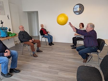Man sieht Senior:innen in einem Stuhlkreis im Aktivhaus der Ambulanten Pflege des DRK in Einbeck. Ein Herr wirft einer anderen Person im Kreis einen gelben Ball zu