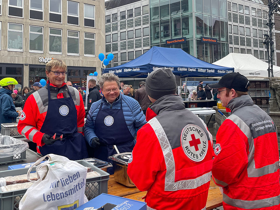 Männer mit DRK-Jacken verteilen am Stand auf dem Marktplatz Erbsensuppen