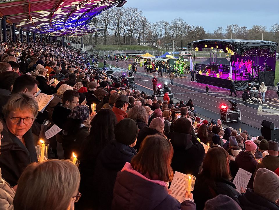 Man sieht die Ränge des Jahnstadions in Göttingen mit vielen Menschen, die zum Adventssingen dort sind. Im Hintergrund sieht man eine Bühne mit lila Beleuchtung
