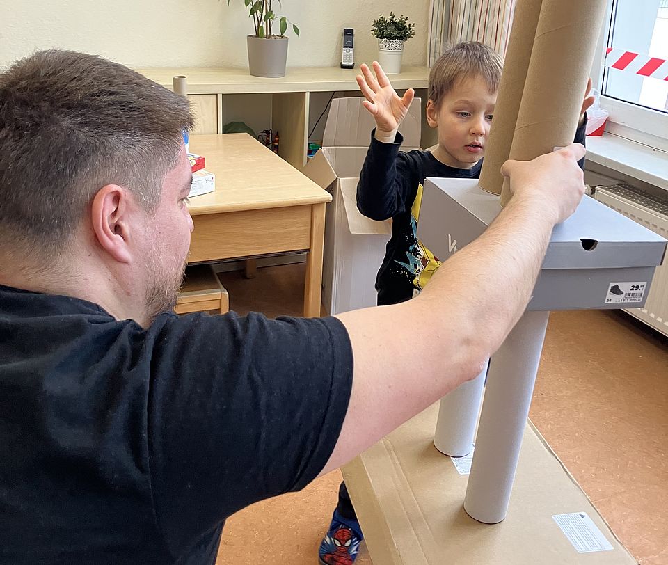 Eine erwachsene Begleitperson und ein Kind bauen gemeinsam einen hohen Turm aus Papprollen, Kartons und weiteren Recyclingmaterialien. Der Turm steht auf einer stabilen Grundplatte und besteht aus mehreren hohen Röhren, die mit einer Schuhschachtel abgestützt werden. Die erwachsene Person hält den Turm seitlich fest, während das Kind gegenüber steht und eine Hand hebt, als würde eine nächste Bauteil‑Entscheidung abgestimmt. Hinter beiden steht ein Regal mit Spielmaterialien und eine Pflanze. Die Szene gehört zum Turmbau‑Wettbewerb der Kinder‑Olympiade in der DRK-Kita Scheden