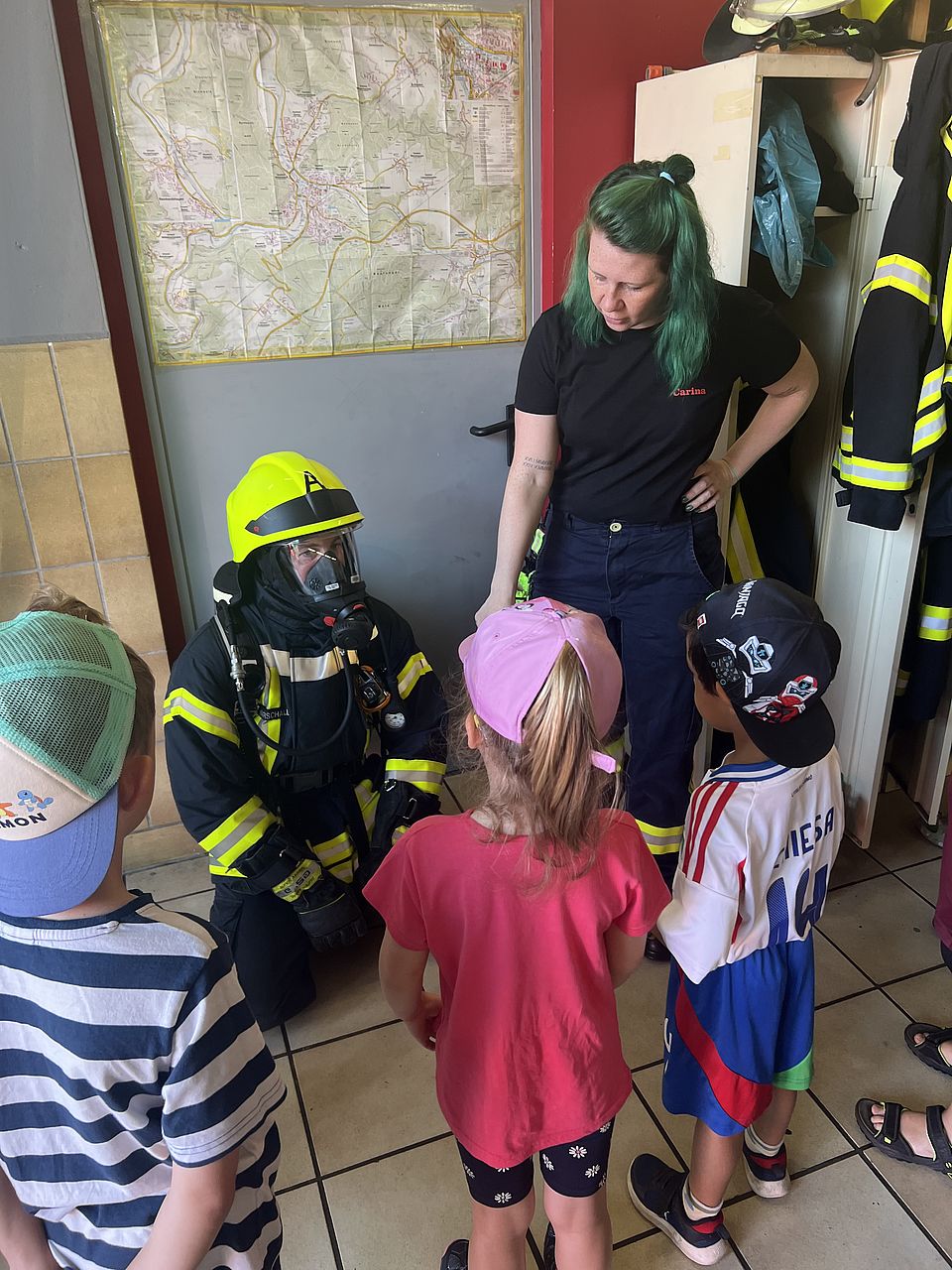 Die Kita-Kinder stehen vor einem Feuerwehrmann, der mit Atemmaske und Uniform vor den Kindern kniet und eine Feuerwehrfaru erklärt etwas dazu.
