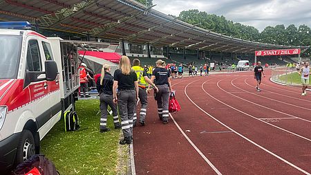 DRK-Bereitschaft beim Sanitätsdienst beim Frühjahrsvolkslauf in Göttingen im Jahnstadion: Bereitschaftsmitglieder in Uniform mit DRK Auto im Stadion, im Hintergrund der Start- und Zielbereich und Rennstrecke