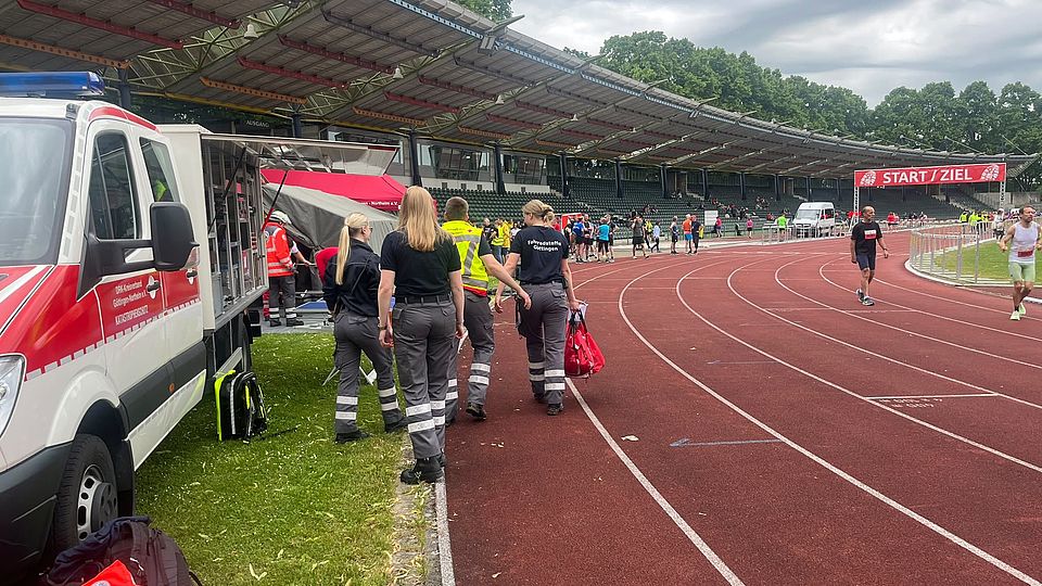 DRK-Bereitschaft beim Sanitätsdienst beim Frühjahrsvolkslauf in Göttingen im Jahnstadion: Bereitschaftsmitglieder in Uniform mit DRK Auto im Stadion, im Hintergrund der Start- und Zielbereich und Rennstrecke
