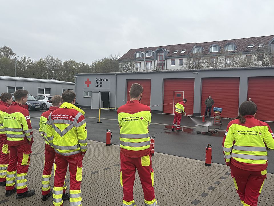 2026_04_13_Brandsschutzübung_im_Rettungsdienst__18_.jpg DRK-Rettungsdienst bei einer Brandschutzübung auf dem Hof des DRK-Kreisverbandes Göttingen-Northeim: Ein Mitarbeitender des Rettungsdienstes in Uniform löscht ein Feuer auf dem Boden, im Vordergrund warten andere Mitarbeitende im Halbkreis und schauen zu