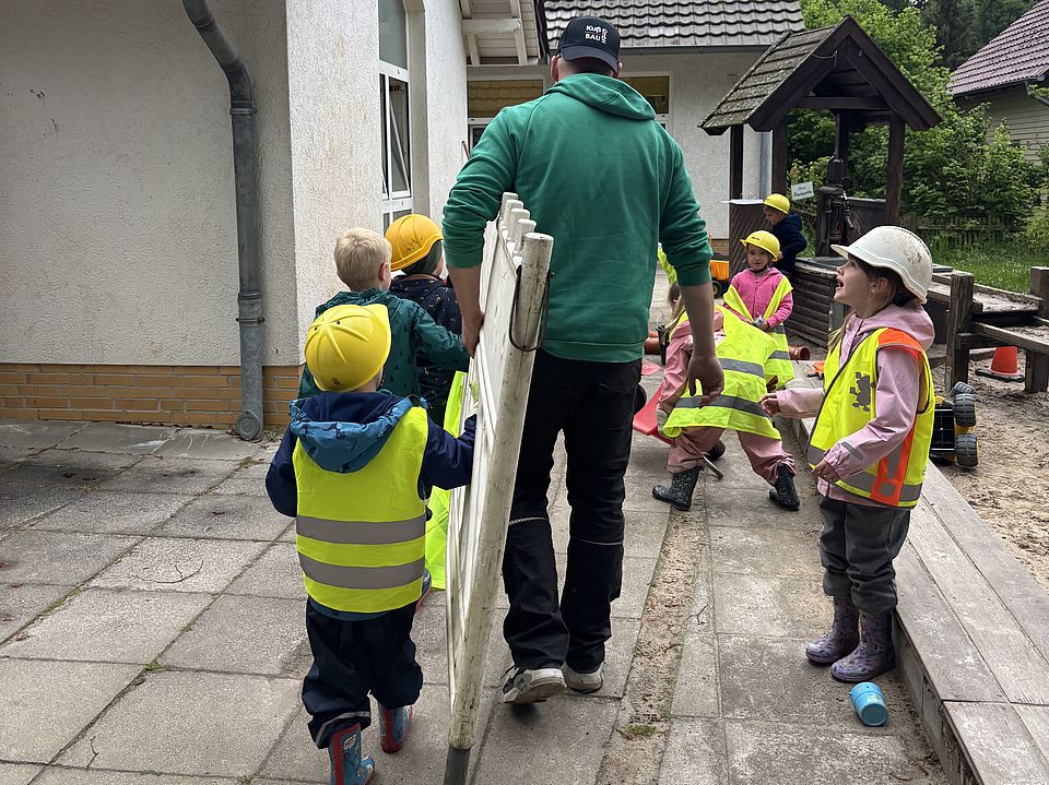 2026_05_27_Baustellentag_für_die_Kinder__5_.jpeg Auf dem Außengelände der Kita trägt eine erwachsene Person eine Leiter, während mehrere Kinder in Warnwesten und Bauhelmen daneben stehen und zusehen. Im Hintergrund sind Spielbereiche und Sandfläche zu sehen.