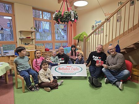 Eine Gruppe von Kindern sitzen mit drei Männern auf einem grünen Teppich in einem Raum der Kita Bovenden. Einer der Männer ist der Leiter der Kita. Er hält gemeinsam mit einem der anderen Männer einen Gutschein der Fa. Freckmann "Sport 2000" in den Händen. DIe beiden anderen Männer tragen ein Sweat-Shirt mit dem Aufdruck "Brutal regional" mit einem roten Herz zwischen den Wörtern und halten ein Schild des Hofladen Zapfe als Werbeschild in die Runde.