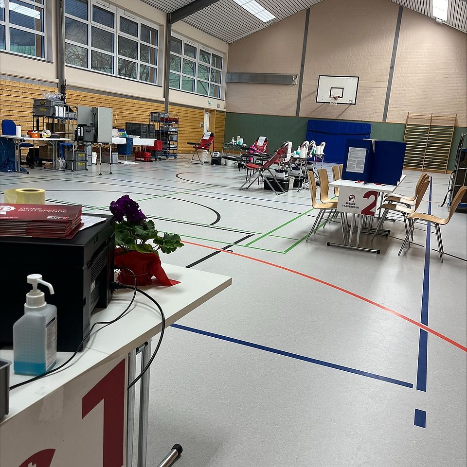 2026_02_Blutspende_Groß_Lengden__4_.jpeg Eine große Sporthalle ist für einen Blutspendetermin vorbereitet. Im Vordergrund steht ein Tisch mit einem Drucker, Informationsmaterialien, einem Desinfektionsspender und einer Blume. Im Hintergrund stehen mehrere Blutspendeliegen mit weiß‑roten Auflagen, eine stationäre Untersuchungsecke sowie Tische und Stühle für den Ablauf der Spende. An den Hallenwänden befinden sich Klettergeräte, Sportmarkierungen sind auf dem Boden erkennbar. Die Szene zeigt den vollständig vorbereiteten Ablaufbereich einer Blutspendeaktion in einer Mehrzweckhalle.