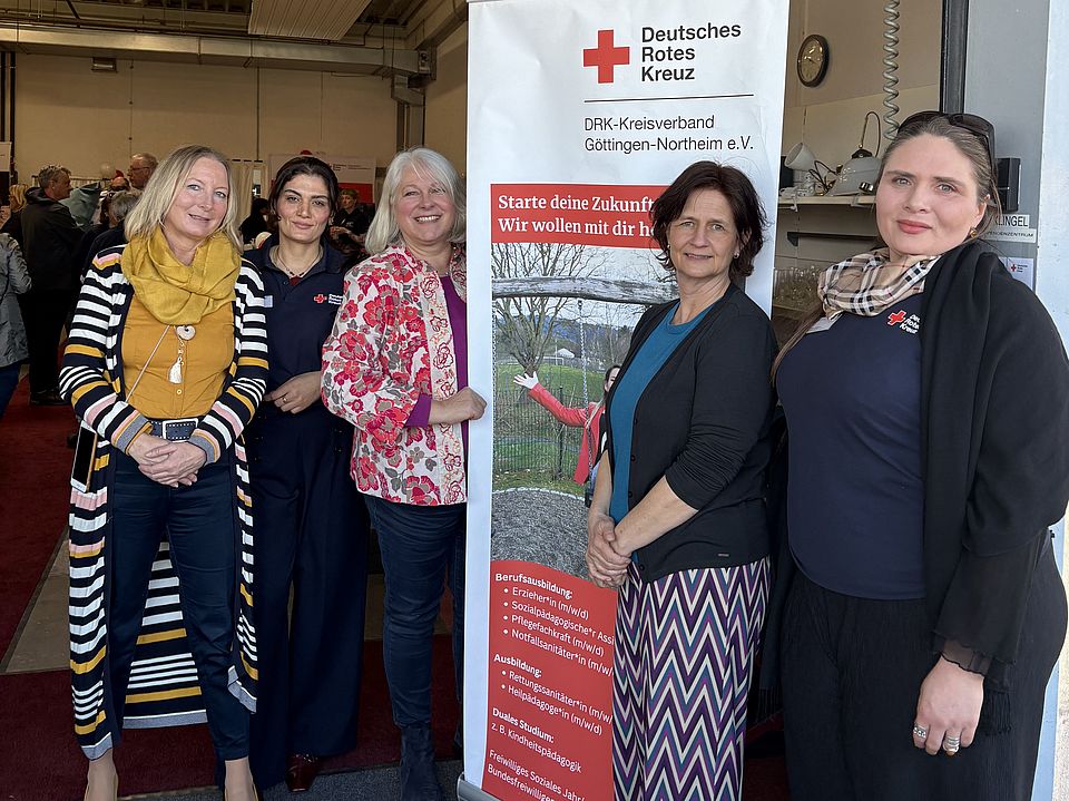 2026_03_03_Spendenzentrum_Einweihung_Frau_Allweil__Frau_Arif__Frau_Krause__Frau_Reußner__Frau_Jablonski Annette Allweil, Nawal Arif, Anja Krause, Petra Reußner, Kinga Jablonski stehen beim Eingang des neuen DRK‑Spendenzentrums Göttingen vor einem DRK‑Roll‑up. Im Hintergrund sieht man zahlreiche Gäste der Einweihungsfeier des ‘Kaufhaus für alle’