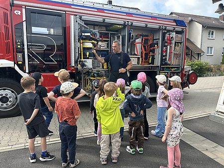 Kinder der DRK-Kita Volkmarshausen stehen vor einem Feuerwehrfahrzeug mit geöffneten Geräteraumklappen, während eine Einsatzkraft die Technik erklärt.