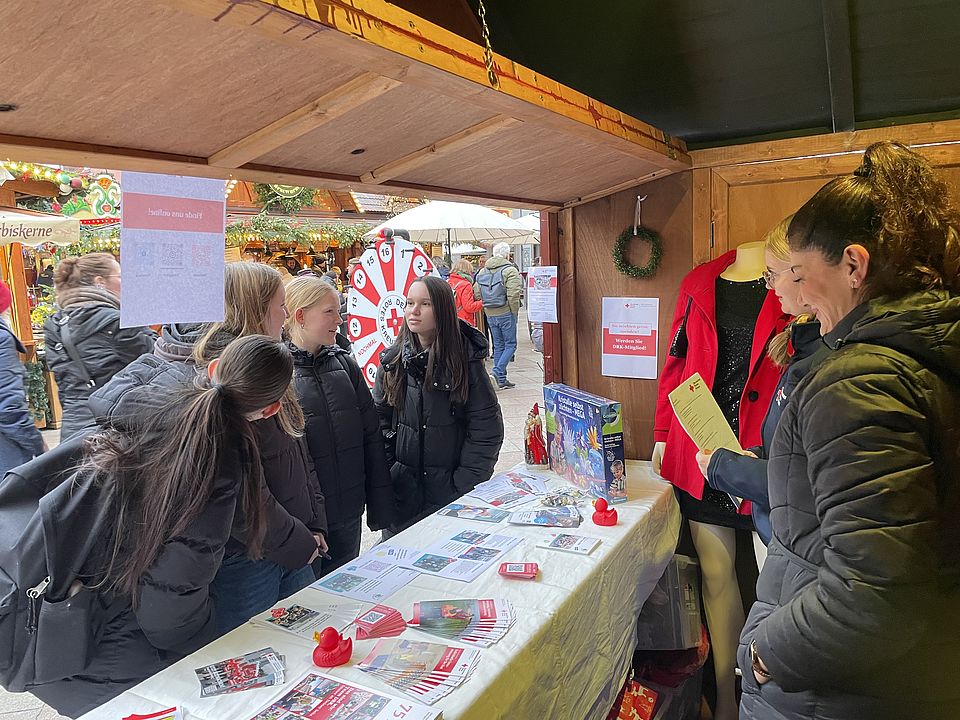 Freiwillig-in-Göttingen-Hütte am 5.12.25 auf dem Weihnachtsmarkt Die Freiwillig-in-Göttingen-Hütte von innen mit zwei DRK Ehrenamtlichen und vier Interessierten, die sich am Stand informieren und das Glücksrad-Quiz beantworten