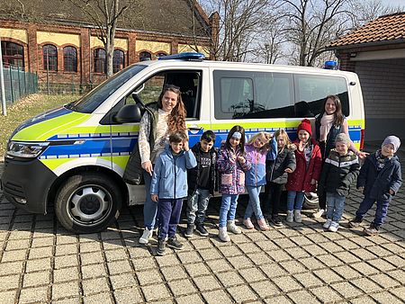 Eine Gruppe Kinder aus der DRK-Kita Scheden vor einem Polizeiauto, gemeinsam mit einer Kita-Mitarbeiterin, bei ihrem Besuch in der Polizeischule
