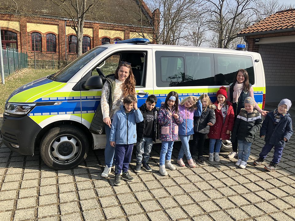Eine Gruppe Kinder aus der DRK-Kita Scheden vor einem Polizeiauto, gemeinsam mit einer Kita-Mitarbeiterin, bei ihrem Besuch in der Polizeischule