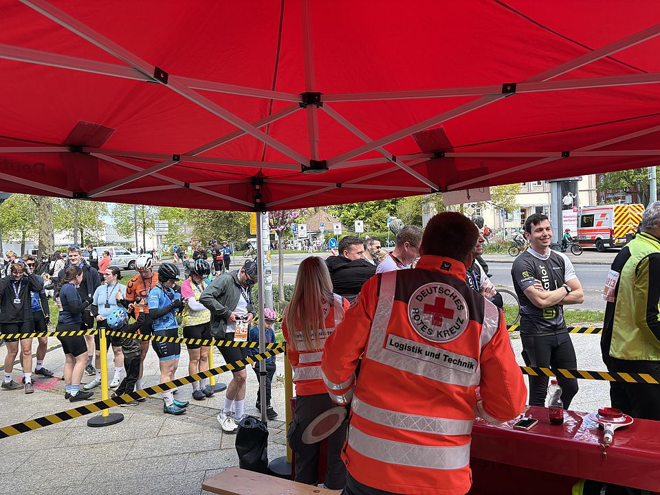 Helfer*innen und Mitglieder der DRK-Bereitschaft bei der Tour d'Energie 2026 in Göttingen beim Verpflegungsdienst, im Vordergrund Mitarbeitende mit orangenen Jacken mit der Aufschrift 'Deutsches Rotes Kreuz'. Eine Schlange an Radfahrer*innen steht an.