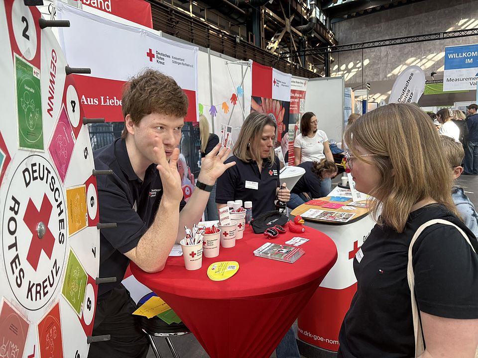 Infotisch des DRK‑Kreisverbands Göttingen‑Northeim e. V. beim GöBit 2026 mit Glücksrad und Werbematerialien. Ein DRK‑Mitarbeitender und eine Besucherin stehen am Tisch.